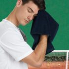 A man in a white shirt wipes his forehead with a dark towel. He stands on a tennis court with a green background and a net visible behind him, appearing to rest after playing tennis.