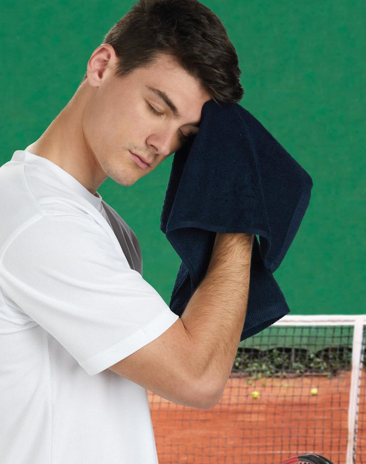 A man in a white shirt wipes his forehead with a dark towel. He stands on a tennis court with a green background and a net visible behind him, appearing to rest after playing tennis.