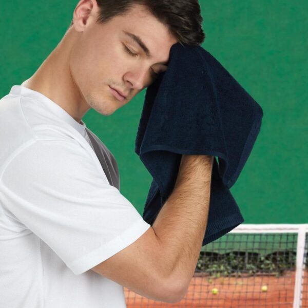A person in a white shirt wipes their forehead with a navy towel, standing beside a tennis court. The court has a net, and tennis balls are scattered on the ground. The background is green.