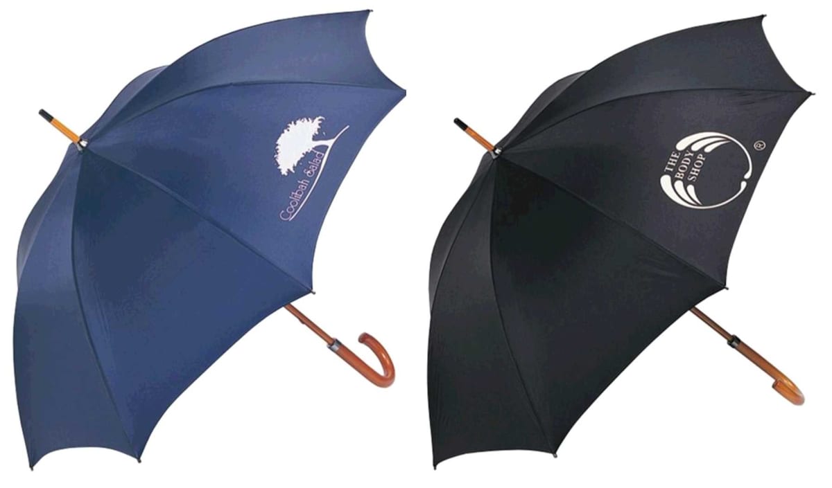 Two large umbrellas with wooden handles are displayed. The left umbrella is navy with a white tree logo and Coastal State text. The right umbrella is black with a logo and text that reads The Body Shop.