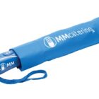 Blue folded umbrella in a matching sleeve, featuring the text MMcatering and a logo on its handle. A wrist strap is attached to the sleeve.