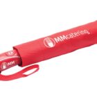 A compact red umbrella with a matching cover. The cover has a logo featuring a fork and circular shape, accompanied by the words MM Catering printed in white. The handle is red with a white logo and includes a wrist strap.