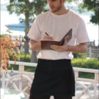 A person in a white t-shirt and black apron stands on a sunny outdoor patio, holding a notebook and pen, appearing to take an order. Greenery and a blurred view of water are in the background. The word HOSPITALITY is visible at the bottom.