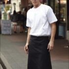 A person in a white T-shirt and black apron stands confidently on a city sidewalk, with a blurred background of buildings and pedestrians. The word HOSPITALITY is written at the bottom of the image.