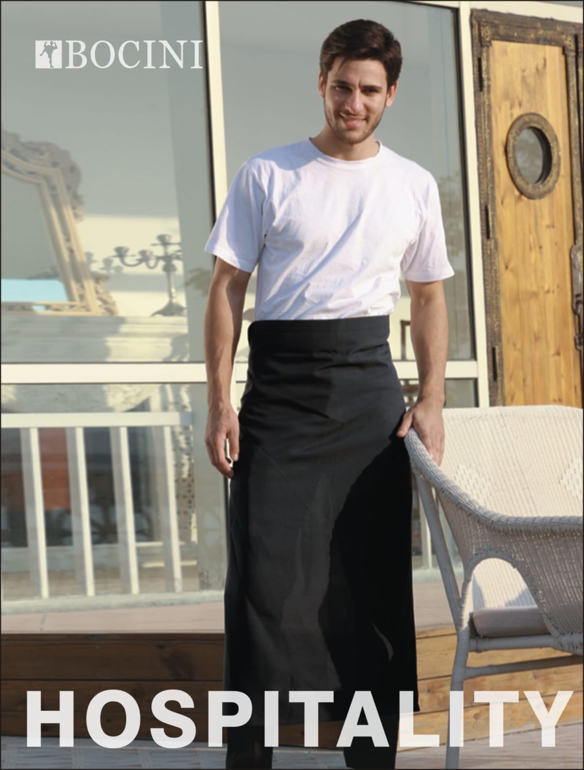A person stands outside wearing a white shirt and a black apron, leaning against a wicker chair. The word HOSPITALITY is displayed at the bottom. The logo BOCINI is at the top left. Theres wooden decor and glass railing in the background.