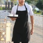 A person in a black apron carries a tray with two white cups outdoors. Theres greenery in the background and a small cart with a bottle next to them. The word HOSPITALITY is written at the bottom.