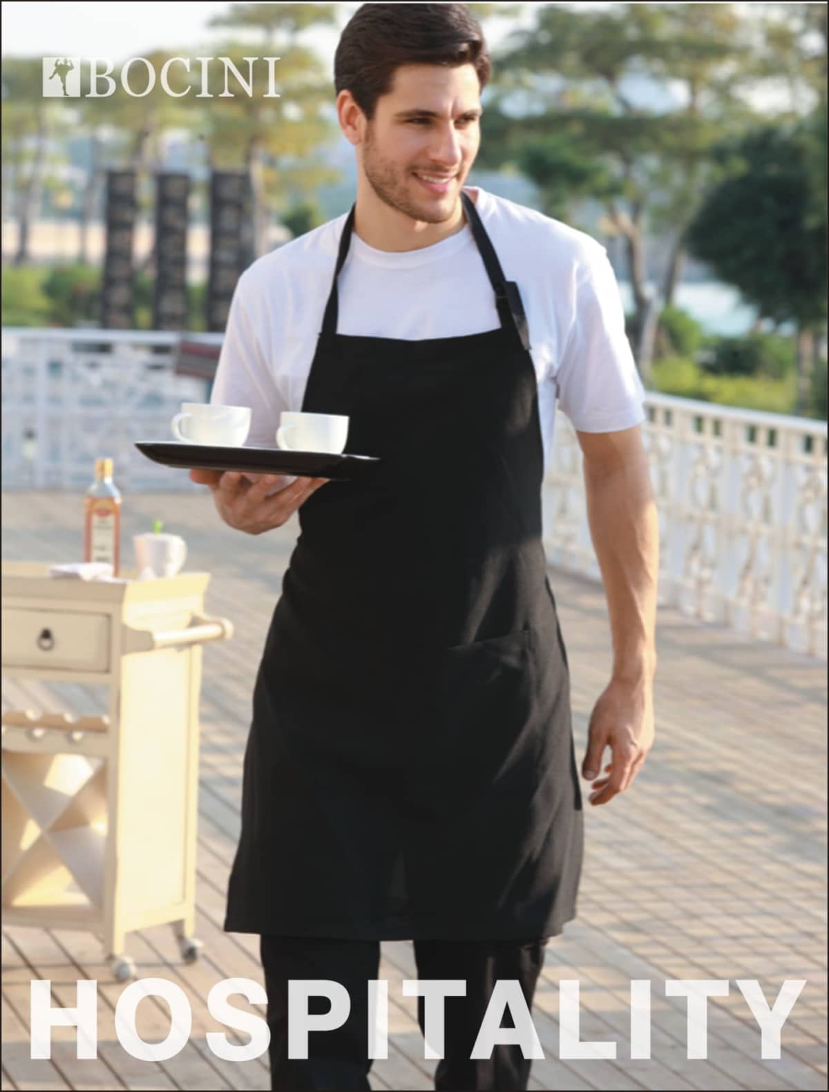 A person in a black apron carries a tray with two white cups outdoors. Theres greenery in the background and a small cart with a bottle next to them. The word HOSPITALITY is written at the bottom.