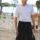 A person in a white t-shirt and black apron smiles while standing outdoors. The background features a railing, trees, and water. The word HOSPITALITY is displayed at the bottom of the image.