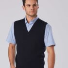 A person wearing a blue short-sleeve collared shirt and a black V-neck vest stands against a plain background, looking to the side with hands by their sides.