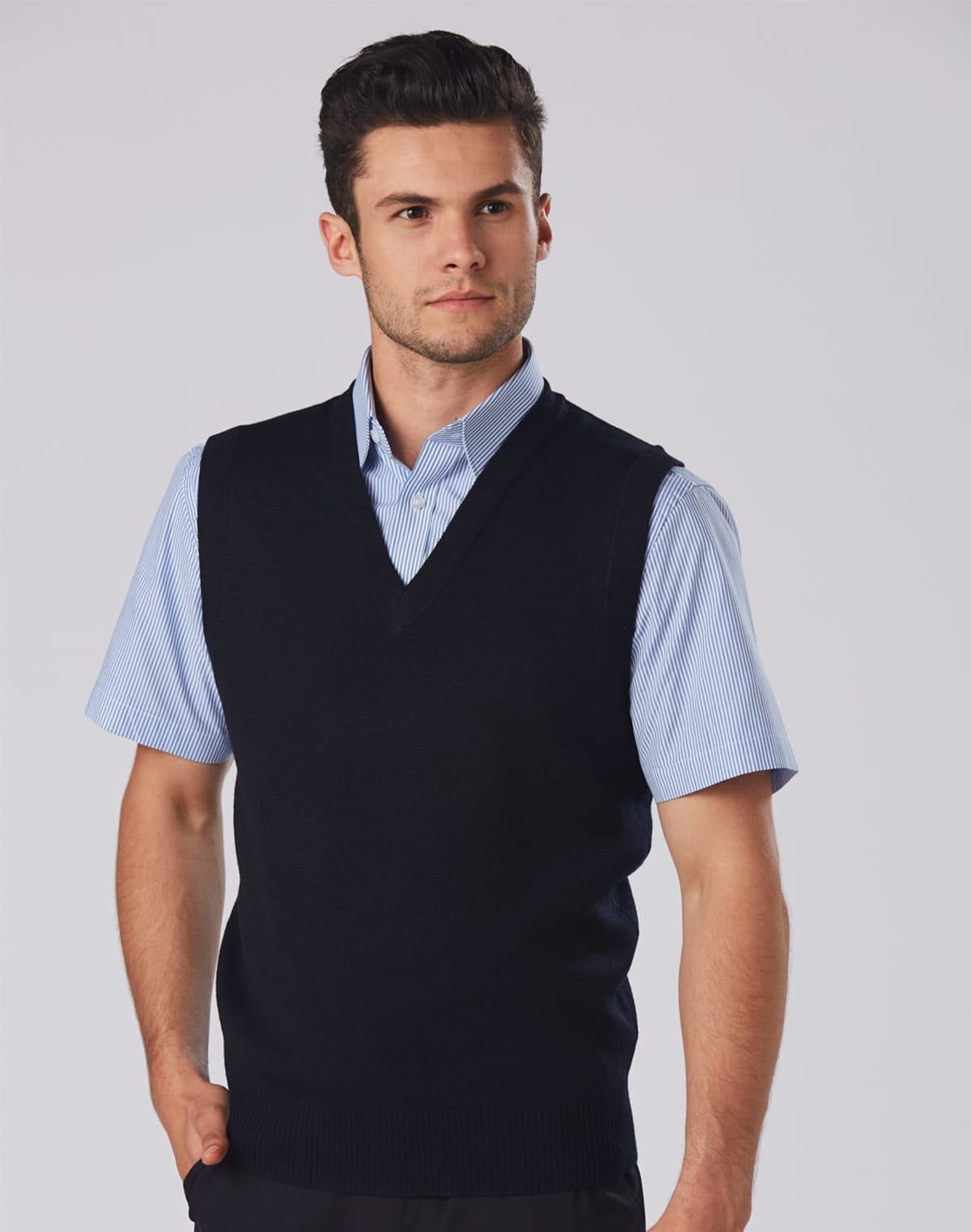 A person wearing a blue short-sleeve collared shirt and a black V-neck vest stands against a plain background, looking to the side with hands by their sides.