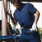 A man in a blue work uniform operates machinery while holding a chain. He wears white gloves and works in an outdoor setting with a car visible in the background. The word WORK WEAR is at the bottom of the image.