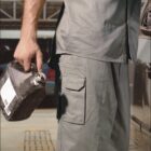 A person wearing gray workwear holds a dark container next to a vehicle. The image has the text BOCINI at the top and WORK WEAR at the bottom. The setting appears to be an industrial or garage area.