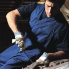 A person wearing a blue work uniform and gloves is focused on repairing a vehicle engine. The word BOCINI is at the top, and WORK WEAR is at the bottom of the image.