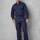 A man stands against a gray background, wearing a navy blue uniform with long sleeves and matching pants. He is dressed in tan boots and has a relaxed posture, with his right hand in his pocket.