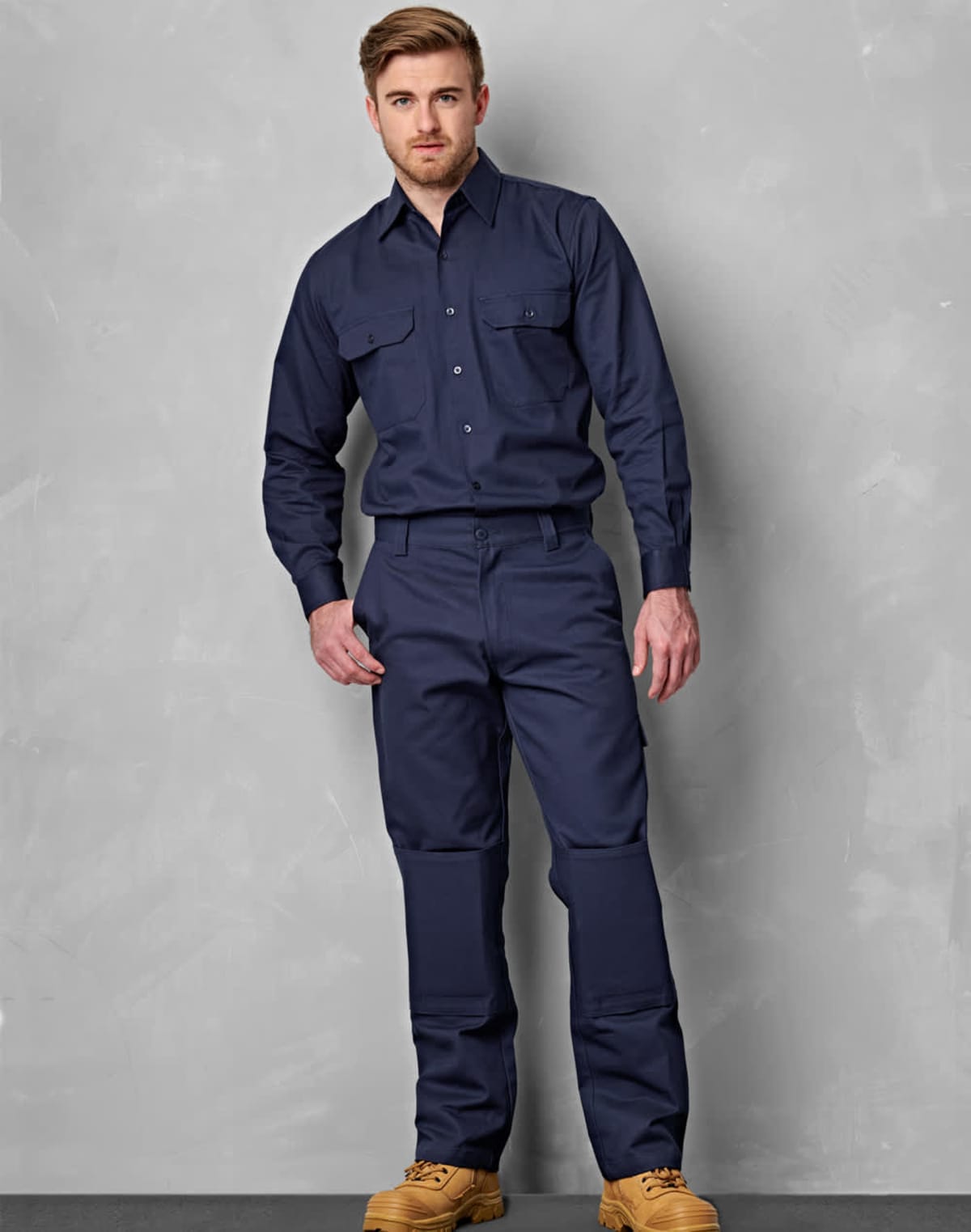 A man stands against a gray background, wearing a navy blue uniform with long sleeves and matching pants. He is dressed in tan boots and has a relaxed posture, with his right hand in his pocket.