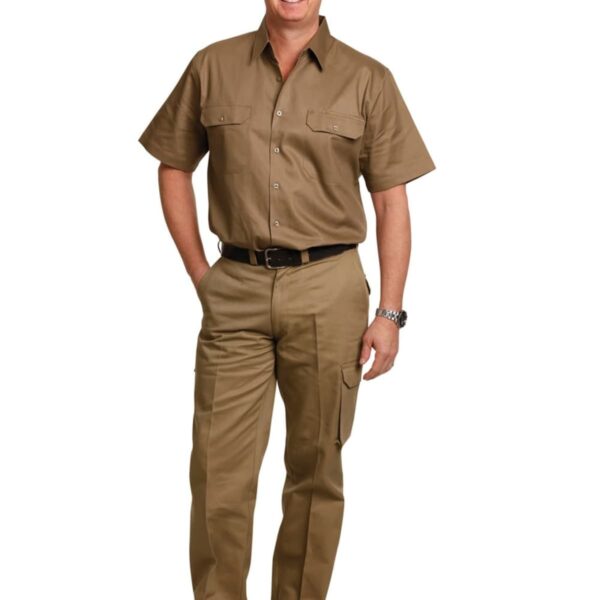 A man smiling, wearing a khaki short-sleeve button-up shirt and matching pants. He has a black belt and wears black shoes, standing with hands in pockets against a plain white background.
