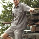A person wearing a gray button-up short-sleeve shirt and matching shorts leans on a stack of wooden beams outdoors. The words WORK WEAR are written at the bottom. Trees are visible in the background.