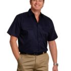 A man smiling, wearing a navy short-sleeve shirt and khaki pants with cargo pockets. He stands with one hand in his pocket against a white background.