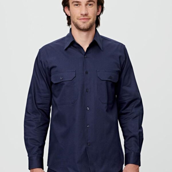 A man with medium-length hair is wearing a dark blue button-up shirt with two front pockets. He stands against a light gray background, looking directly at the camera with a slight smile.