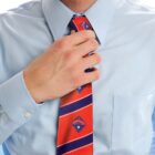 A person in a light blue dress shirt adjusts a red tie with diagonal stripes and small shield emblems. The background is plain white.