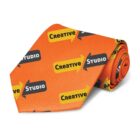 Orange necktie featuring a pattern of yellow and black arrows with the words Creative Studio printed on them. The tie is rolled up to display the design.