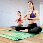 Two people practicing yoga in a bright room, seated cross-legged on green mats with hands in a prayer position. They wear athletic clothing, focusing forward with calm expressions. Light streams in through windows on the right.
