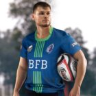 A rugby player in a blue and green jersey holding a rugby ball under one arm. The jersey features logos and the letters BFB. The background is blurred, showing greenery.