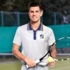 A tennis player stands on a court holding a racket and a tennis ball. He is wearing a white polo shirt with a logo, and there is a net and green fencing in the background.