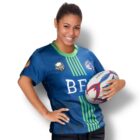 A woman smiles while holding a rugby ball under her arm. She is wearing a blue rugby jersey with green stripes, and logos are visible on the shirt. Her hair is tied back, and she stands against a white background.