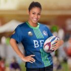 A woman in a blue and green rugby jersey holds a rugby ball, smiling confidently. She stands outdoors on a sunny day with blurred people and tents in the background.