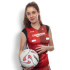 A woman in a red and black sleeveless jersey with Champions text, holding a volleyball. She has long hair tied back, standing against a plain white background and smiling at the camera.