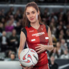 A volleyball player in a red and black jersey labeled Champions holds a volleyball on an indoor court, with a blurred crowd in the background.