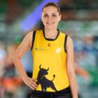 A person wearing a yellow sports jersey with a black bull graphic on the front, standing outdoors with a hand on their hip. The jersey also has logos and a number on the upper front. The background is blurred with hints of green and orange.
