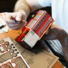 A person assembles a model fire truck made of wood. The truck is red and sits on a table next to sheets of unassembled wooden parts. The person holds a small wheel in one hand.