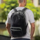 A person wearing a light gray t-shirt and a large black backpack stands outdoors. The background features blurred green trees and buildings, suggesting an urban setting. The image focuses on the back view of the person.
