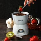 A hand dips a strawberry into a chocolate fondue pot, surrounded by fruit and marshmallows.