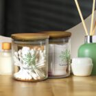 Two glass jars with wood lids sit on a wooden surface. One jar contains cotton swabs, and the other holds cotton pads. Both jars feature the NatureLife logo. In the background are a small bottle, a container, and a diffuser with reeds.