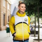 A woman stands outdoors wearing a bright yellow and white jacket with a geometric pattern and a logo. She has her hands in the jacket pockets and is smiling. The background shows a tree and an urban setting with blurred figures.