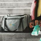 Person tying teal sneakers next to a gray PLEXWELL gym bag on concrete steps.