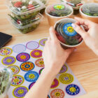 Hands placing a bowl of salad with a smiley face sticker on a wooden table. Surrounding are other bowls, colorful stickers with positive messages like Be Happy, and stacked salads in clear containers.