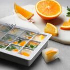 Freezer tray with orange slices and herbs, next to sliced oranges on a cutting board.