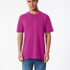 A person wearing a vibrant magenta T-shirt and blue jeans stands against a plain, light background. They have short hair and a neutral expression, looking directly at the camera.