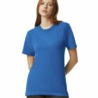 A person with long red hair wearing a blue t-shirt and dark pants stands against a white background. They have a neutral expression and their hands are behind their back.