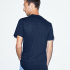 Man wearing a navy blue T-shirt and light blue jeans is standing and facing slightly away from the camera against a plain white background.