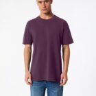 A person stands against a plain white background wearing a plain purple T-shirt and blue jeans. They have short hair and are looking towards the camera with a neutral expression.