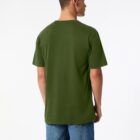 A person with short hair is standing and looking to the left, wearing a green T-shirt and blue jeans. The background is plain white.