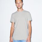 A man stands smiling in a light gray t-shirt and blue jeans against a plain white background. His hair is short and dark, and his hands are relaxed by his sides.