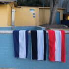 Three striped towels (blue, black, red) hang on a tiled poolside wall near a restroom sign.