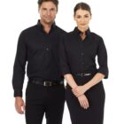 A man and a woman are standing together, both wearing black button-up shirts. The man has his hands by his sides, and the woman is smiling with her hands clasped in front. Both are dressed in a professional manner.