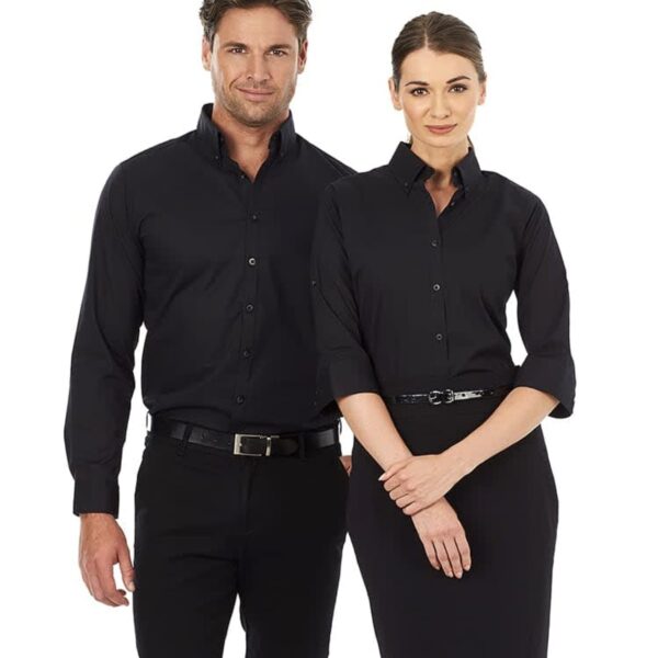 A man and a woman stand side by side, both wearing matching black button-up shirts and black pants. The man has his hands by his sides, while the woman has her hands gently clasped in front. They are smiling at the camera.